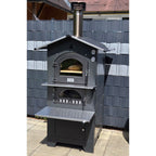 Holzbackofen Gusto Fontana 80x45 freistehend im Garten platziert vor einem klassischen Gartenzaun, ideal für den Außenbereich.