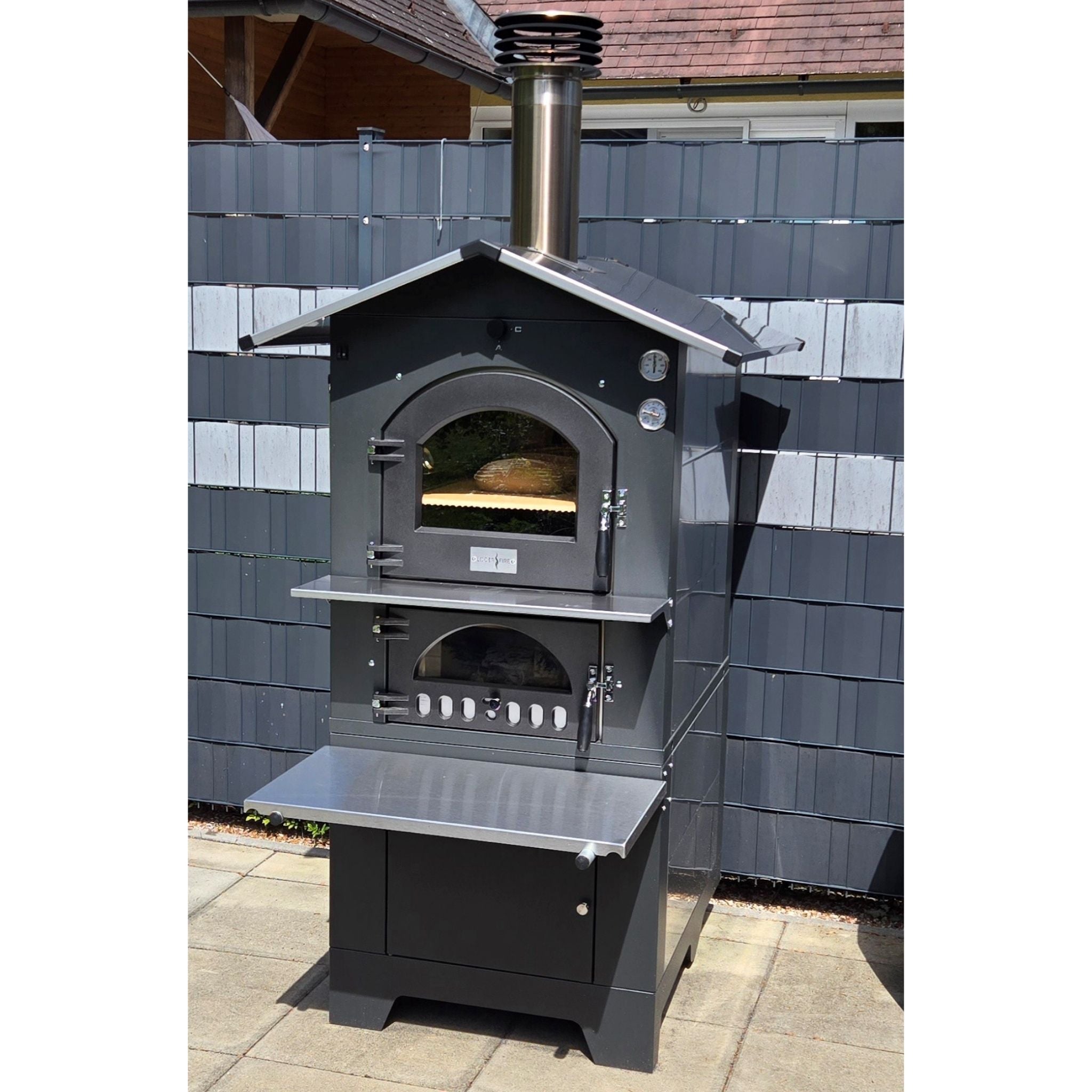 Holzbackofen Gusto Fontana 80x45 freistehend im Garten platziert vor einem klassischen Gartenzaun, ideal für den Außenbereich.